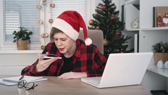 A Cheerful Teenage Boy in a Santa Hat Chatting on a Mobile Phone Sitting at a Computer Table Indoors