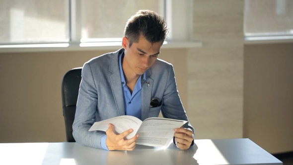 A Young Man In a Suit Jacket Running Notary Flipping Through Documents In Office With a Table On
