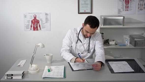 Young Serious Doctor Sitting In Medical Office With Folder And Reading Documents