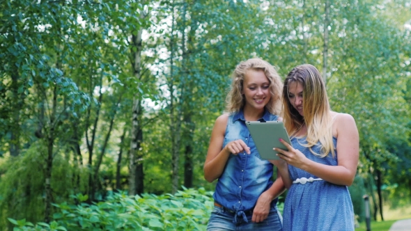 Two Young Woman Walking In The Park, Laughing: Holding The Tablet In Hands
