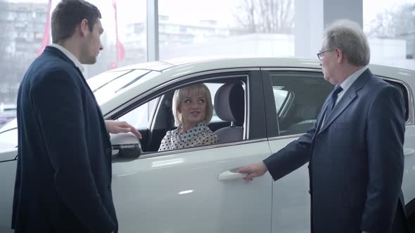 Two Caucasian Men Standing Next To New Car in Dealership As Beautiful Mature Woman Sitting Inside