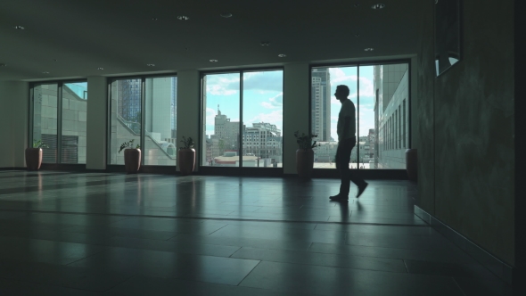 A Man In a Light Shirt Slowly Goes Dark Corridor Of Office Space With Tall Windows.