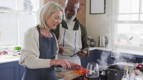 Mixed race senior couple wearing aprons using digital tablet while cooking in the kitchen at home