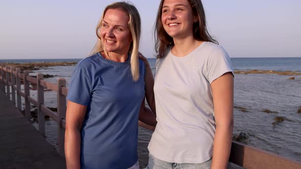 Mother and daughter standing on sidewalk by the sea at sunset