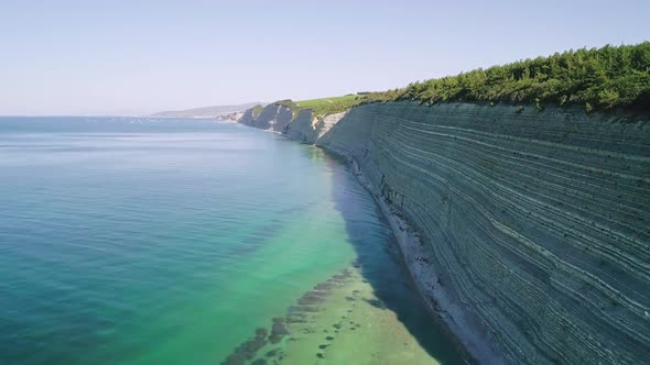 Aerial of High Steep Cliffs Covered with Pine Trees and Wild Pebble Beach with Turquoise Sea Water