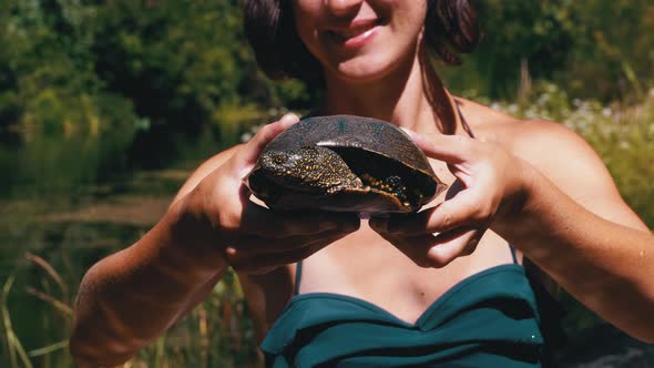 Woman Holds Funny Turtle in Arm and Smiles on River with Green Vegetation