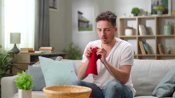 Young Handsome Man Knitting Red Scarf with Knitting Needles While Sitting on Sofa in His Living Room
