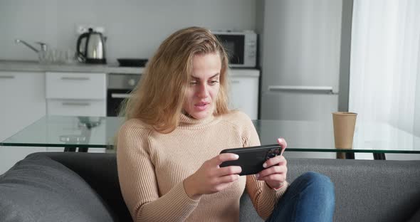 Blonde Millennial Woman in a Beige Woolen Sweater Play Game with Smart Phone on Modern Kitchen