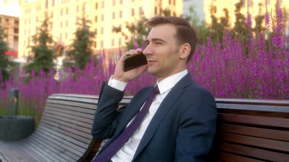 Close Up Young Busy Businessman Sits on Bench with Purple Flowers in Flower Beds and Use Mobile