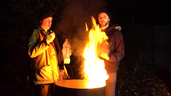 Two Homeless Young Men Stand By the Fire and Eat Apples. Men Stand at Night Near a Barrel of Fire