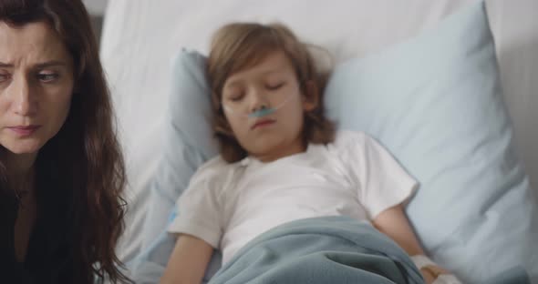 Sick Kid Boy with Lying in Bed at Hospital and Mother Holding His Hand