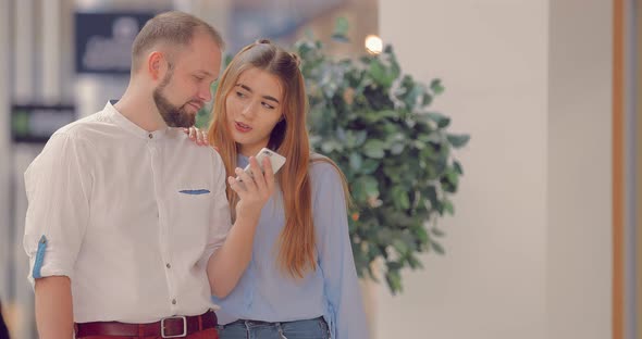 Man and a Woman in a Mall are Looking at a Phone and Talking About Something