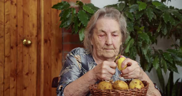 a Purposeful Elderly Woman Sits in Her House and Peels Potatoes That are in a Basket