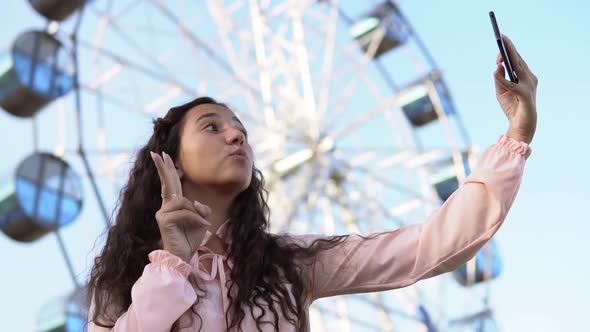 a Girl with Long Hair in a Dress Makes Selfie Using a Phone Standing Near the Ferris Wheel