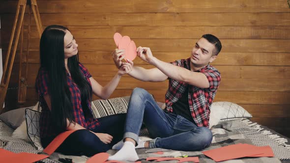 Enamored are sitting on the bed and holding red paper hearts in their hands and playing.