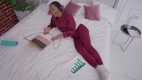 Top View of Joyful Slim African American Young Woman Lying on Bed Conferencing Online on Laptop