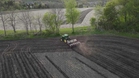 Aerial view of farmer fertilizing and seeding field in straight rows. Trees and hillside.