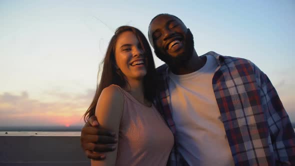 Cheerful Multiracial Couple Hugging and Smiling at Camera Against Sunset View