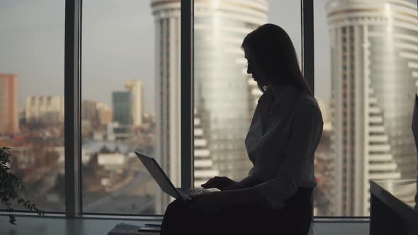 Silhouette of an Attractive Girl Working on a Laptop in an Office Near the Window. Business Woman