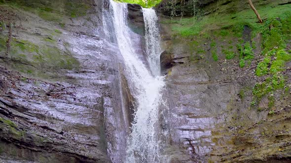 Slow motion tilt down of falling waterfall down green mossy mountain wall into stream of wilderness