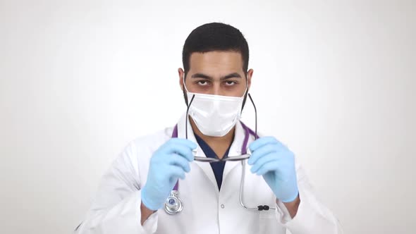 A cheerful Arab male doctor in a lab coat puts on glasses, looks at the camera 