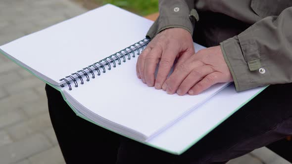 Senior Blind Man Sitting on Bench in City Park and Reading a Braille Book