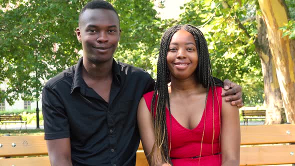 A Young Black Couple Sits on a Bench in a Park and Smiles at the Camera on a Sunny Day
