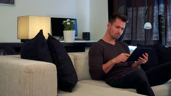 A Young, Handsome Man Sits on a Couch in a Cozy Living Room and Uses His Tablet