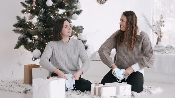 Two Attractive Women Sitting Udr Th Christmas Tree and Throwing Masks in the Air