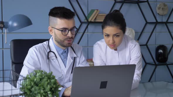 Mixed-races young couple of male and female doctors sitting in hospital, talking and using laptop