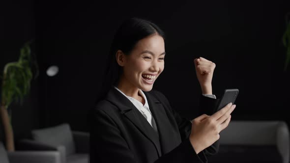 Asian Businesswoman Using Phone Gesturing Yes Celebrating Success In Office