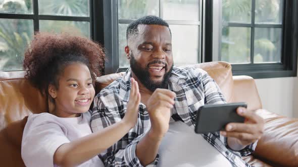 Happy African family dad and daughter having fun and using mobile phone video call on sofa at house