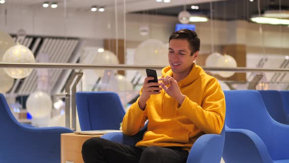 Young Happy Man in Yellow Hoodie Using Smartphone at Cozy Cafe at Evening Time