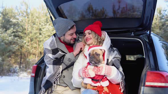 Young Couple a Man and a Woman with Dog are Sitting in the Trunk of a Car in a Winter and Hugging