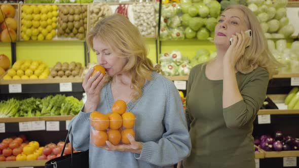 Beautiful Senior Caucasian Woman Smelling Fresh Tasty Tangerine As Her Female Friend Talking on the