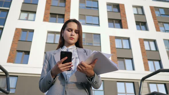 Young Woman in a Business Suit Looks at the Phone and at the Documents on the Background of the