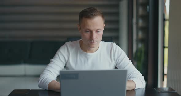 Happy Man Sits in Room at Desktop with Laptop and Rejoices in His Success