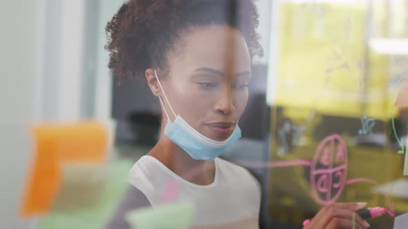 Mixed race businesswoman with face mask writing on board with memo notes in office