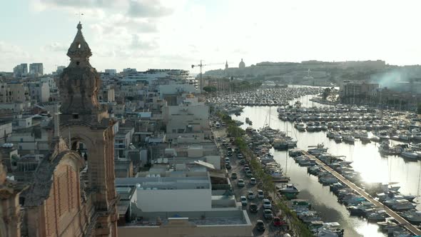 Small Village in Malta Revealing Behind Two Church Bell Towers with Boats in Port on Sunny Day