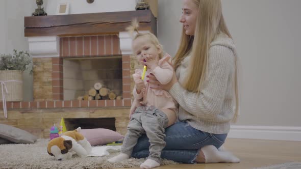 Young Caucasian Woman with Long Hair Putting Jeans on Cute Baby Girl and Taking Party Whistle Off