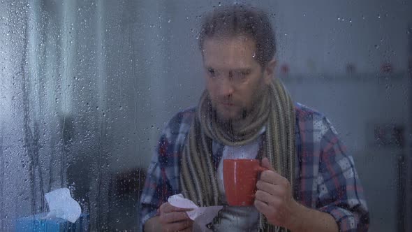 Sick Man Sneezing and Drinking Hot Tea at Home Behind Rainy Window