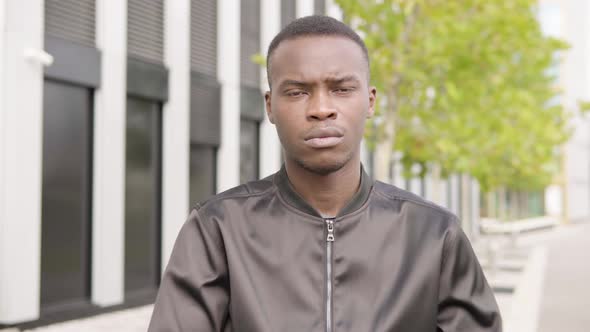 A Young Black Man Looks Seriously at the Camera - an Office Building in the Blurry Background