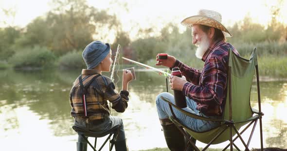 Senior Grandfather and His Cheerful Small Grandson which Fishing on the Pond and Enjoying Hot Tea