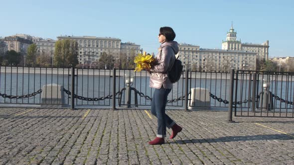 A Middleaged Woman Brunette with a Bouquet of Yellow Leaves Walks Along the City Embankment