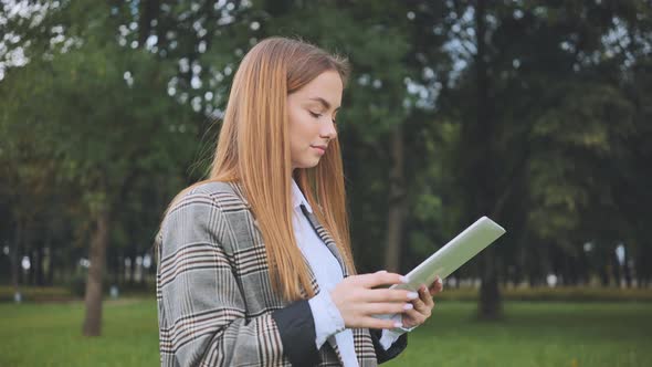 A Young Girl Walks with a Tablet in the Park