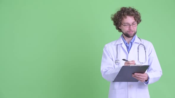 Happy Young Bearded Man Doctor Presenting Something While Writing on Clipboard