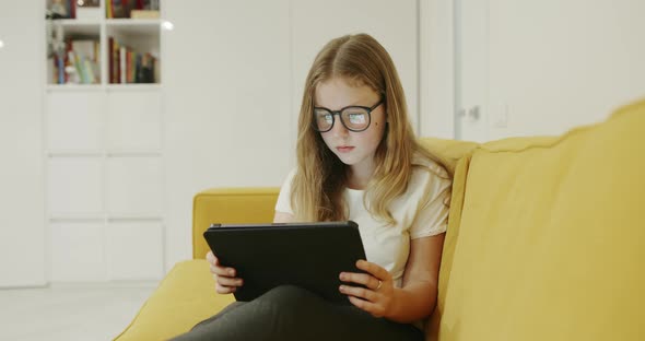 Young Blond Girl Wearing Glasses Sitting on a Comfortable Sofa at Home Using a Tablet-pc During the