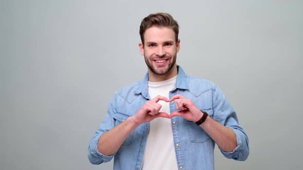 Portrait of handsome young caucasian man making heart sign by hands over grey background