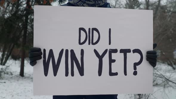 Man stands in a field covered in snow and holds a sign that says did i win yet