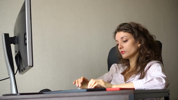 Tired Young Woman with Curly Hair and White Shirt is Working at the Office Using Her Computer